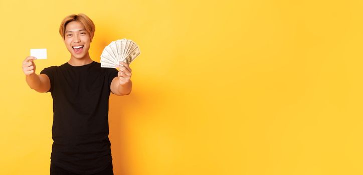 Happy good-looking asian guy in black casual clothes, showing money and credit card, smiling sassy, yellow background.