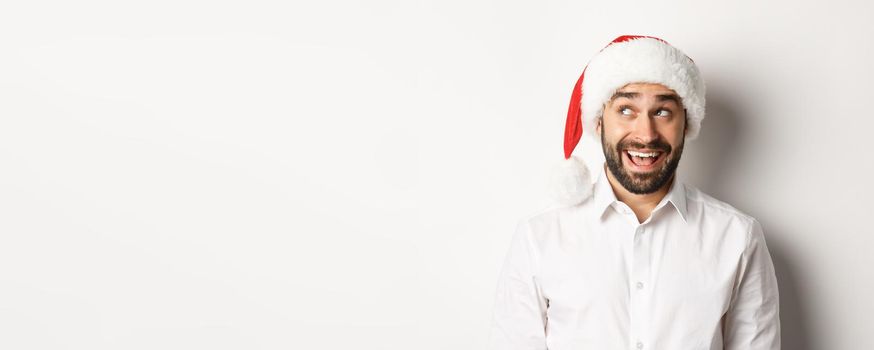 Close-up of happy bearded man in santa hat, celebrating christmas and New Year and looking at upper left corner, imaging something. white background.