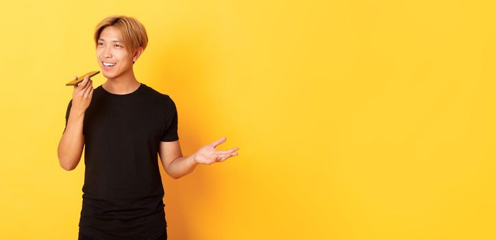 Portrait of stylish handsome korean guy with blond hair record voice message on mobile phone, holding smartphone near mouth and speaking, standing yellow background.