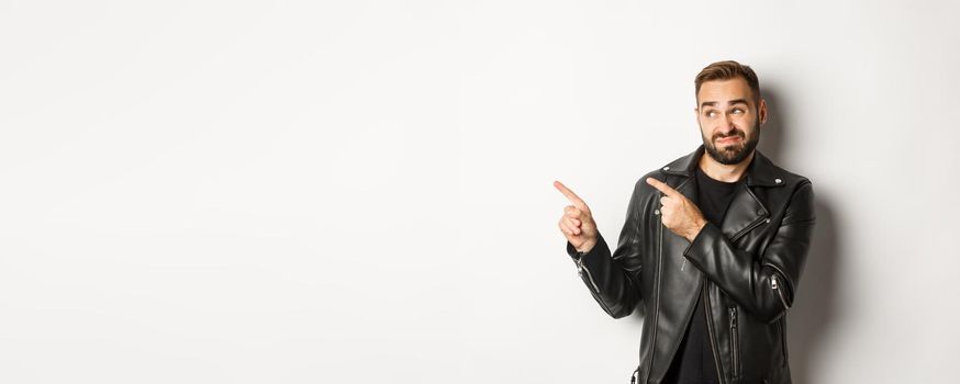 Skeptical and doubtful guy in black leather jacket, shrugging while pointing at upper left corner promo offer, standing over white background.