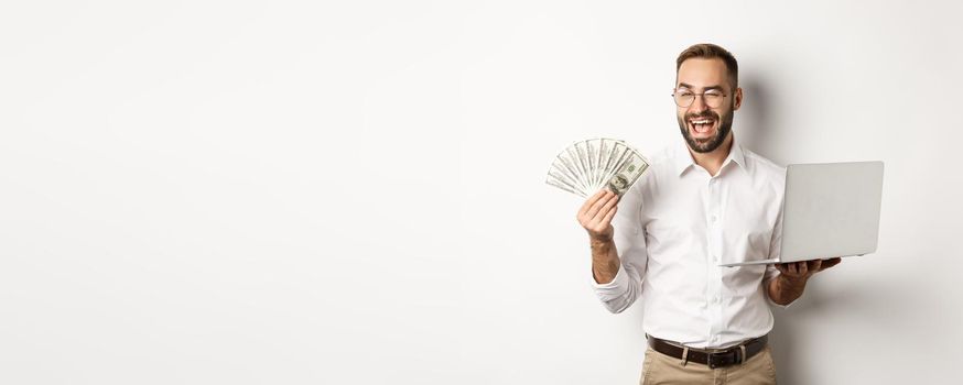 Business and e-commerce. Confident businessman showing how work online, winking, holding money and laptop, standing over white background.