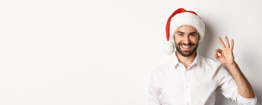 Party, winter holidays and celebration concept. Confident man in santa hat showing okay sign, approve and like, white background.