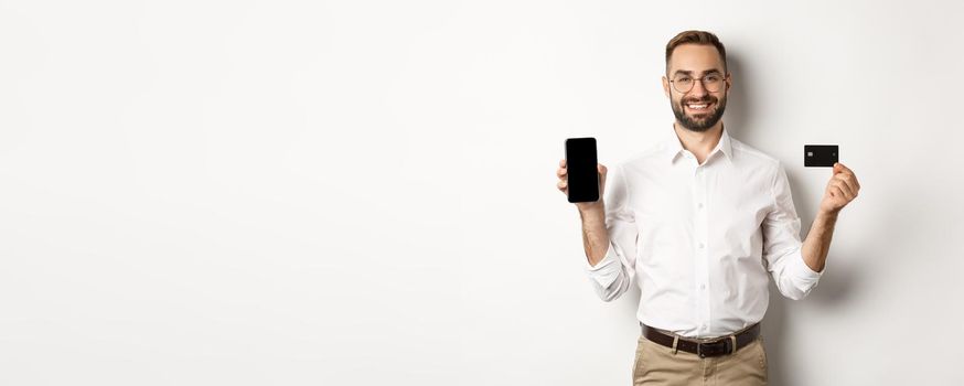 Business and online payment. Smiling handsome man showing mobile screen and credit card, standing over white background.