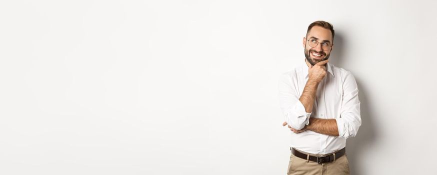 Satisfied businessman having interesting idea, looking at upper left corner and thinking with pleased smile, standing over white background.