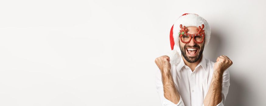 Close-up of man celebrating christmas or new year, wearing xmas party glasses and santa hat, rejoicing and shouting of joy, standing over white background.