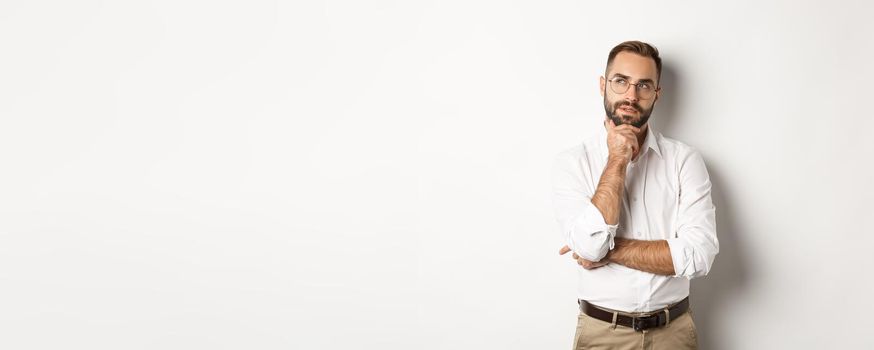 Thoughtful businessman in glasses making plan, looking at upper left corner and thinking, standing against white background.