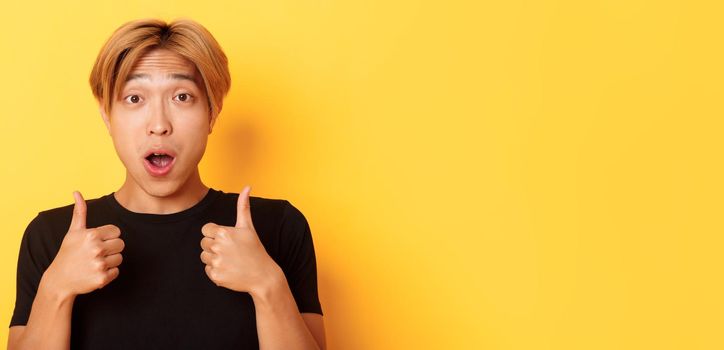 Close-up of impressed and excited handsome asian guy showing thumbs-up and looking amazed, standing over yellow background.