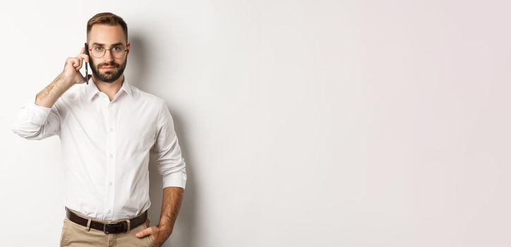 Confident business man talking on phone, looking serious, standing over white background.