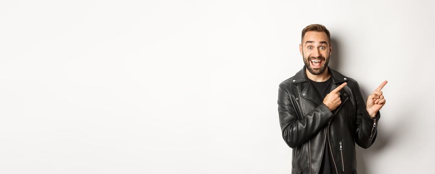 Excited, handsome man in black leather jacket, pointing fingers right at promo offer, showing logo, white background.