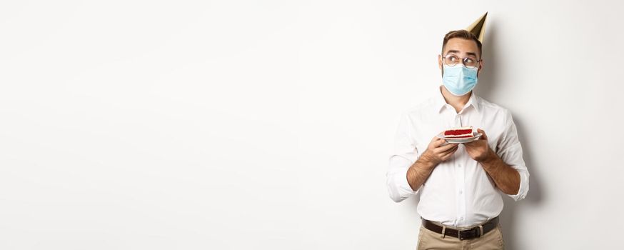 Covid-19, social distancing and celebration. Thoughtful man holding birthday cake, making wish and wearing face mask on quarantine, white background.