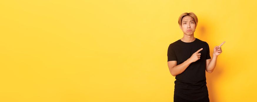 Portrait of disappointed and sad handsome blond asian guy, sulking upset, pointing fingers upper right corner, standing yellow background.