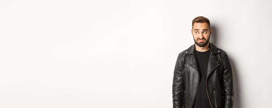 Indecisive gloomy man in black leather jacket, looking left and feeling uncomfortable, standing against white background.