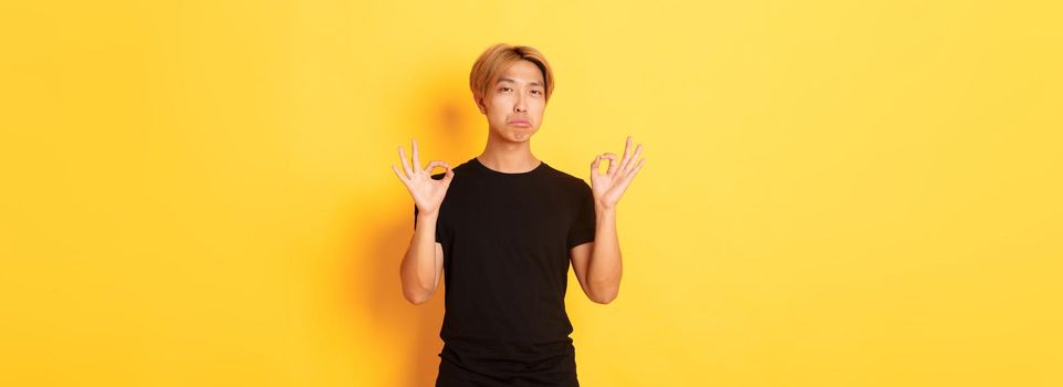 Impressed asian stylish guy with blond hair, showing okay gesture and praise something good, standing yellow background.