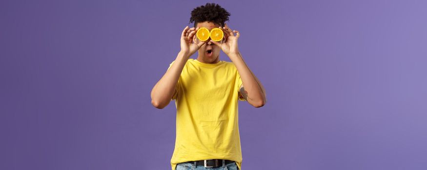 Holidays, vitamins and vacation concept. Portrait of funny, playful young guy fool around, playing with fruits, making eyes from pieces of orange, show wondered face expression, purple background.