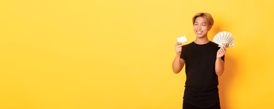Portrait of satisfied smiling, handsome asian guy looking pleased at credit card while also holding money, standing yellow background.