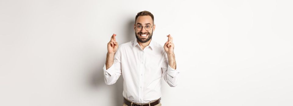 Hopeful businessman smiling and cross fingers for good luck, making a wish, standing over white background.