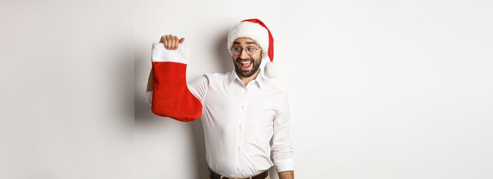 Merry christmas, holidays concept. Happy adult man receive gifts in xmas sock, looking excited, wearing santa hat, white background.