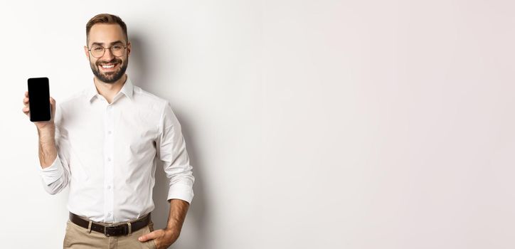 Satisfied business man showing mobile screen, smiling proudly, standing over white background.