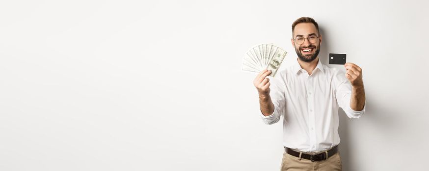 Happy young man showing his credit card and money dollars, smiling satisfied, standing over white background.