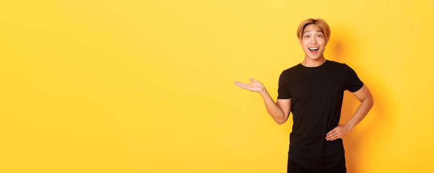 Portrait of happy and excited smiling asian guy holding something on hand over yellow background.