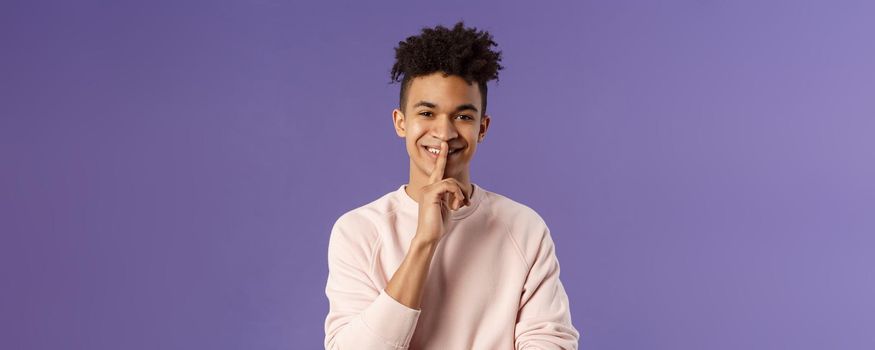 Close-up portrait of attractive smiling young man hiding secret, asking keep silent or quiet to do surprise, show shush gesture, place index finger to mouth, standing purple background.