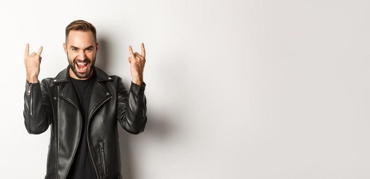 Cool adult man in black leather jacket, showing rock on gesture and tongue, enjoying music festival, standing over white background.