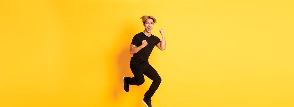 Full-length of happy attractive asian guy in black clothes jumping and celebrating victory, achieve goal, standing yellow background, triumphing.