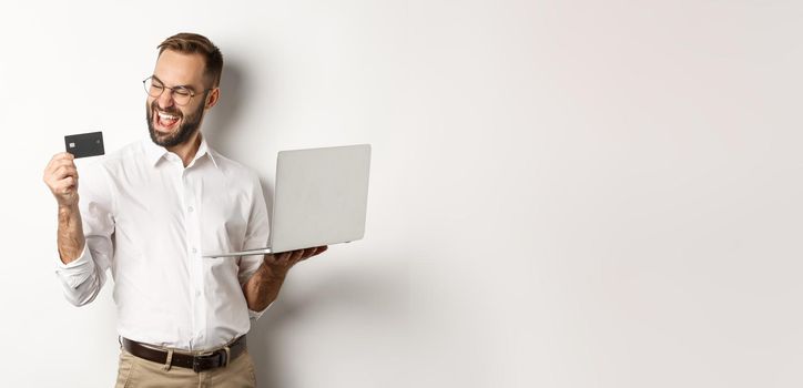 Online shopping. Satisfied handsome man looking at credit card after making order internet, using laptop, standing over white background.