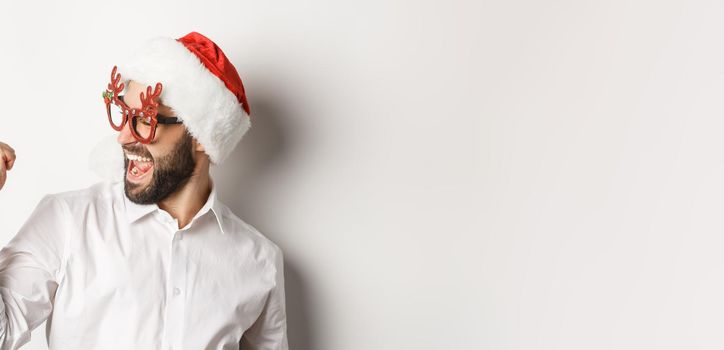 Close-up of happy bearded man wearing santa hat and christmas party glasses, making fist pump and rejoicing, achieve goal and celebrating, white background.