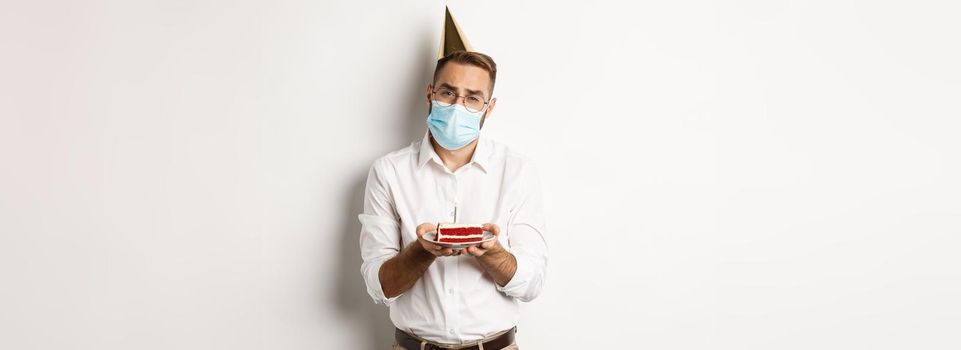 Coronavirus, quarantine and holidays. Sad man cant blow candle off birthday cake, wearing face mask and complaining, standing over white background.