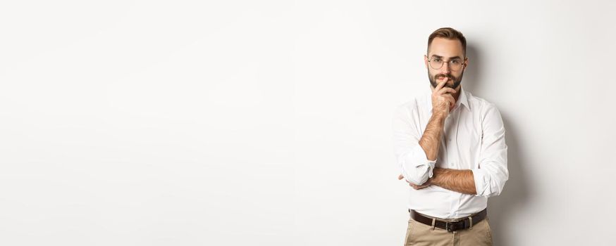 Thoughtful handsome businessman looking at camera, making choice or thinking, standing in glasses and white collar shirt against studio background.