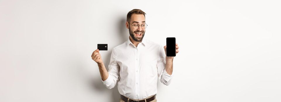 Business and online payment. Smiling handsome man showing mobile screen and credit card, standing over white background.