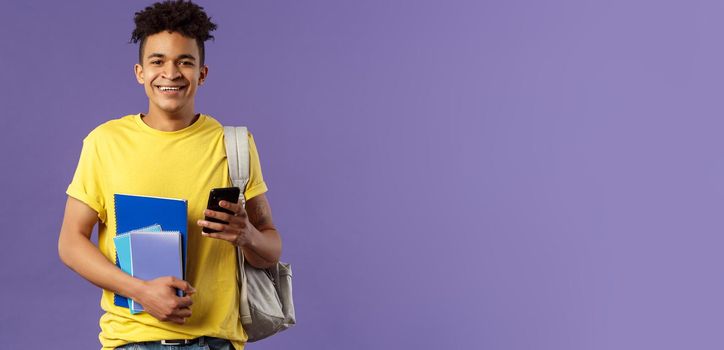 Back to school, university concept. Portrait of cheerful young handsome male student wearing backpack on shoulder, hold notebooks and studying books, texting friend mobile phone.