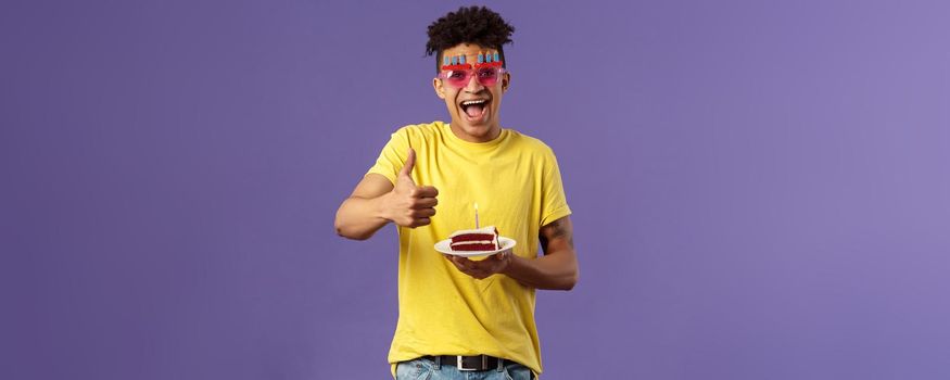 Celebration, party and holidays concept. Portrait of upbeat, cheerful young happy man celebrating birthday, wear funny sunglasses and hold b-day cake with lit candle, show thumb-up, recommend.