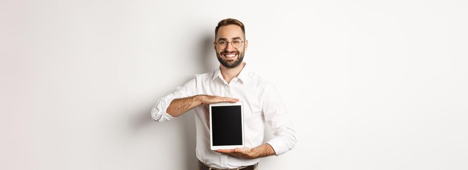 Smiling manager showing something on digital tablet screen, demonstrating website, standing over white background.