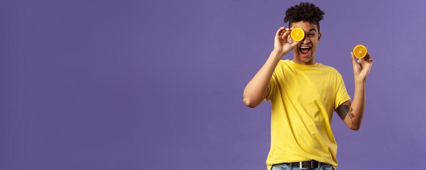 Holidays, vitamins and vacation concept. Portrait of carefree, upbeat good-looking man having fun, look playful laughing, likes eating fruits healthy food, holding pieces of oranges.