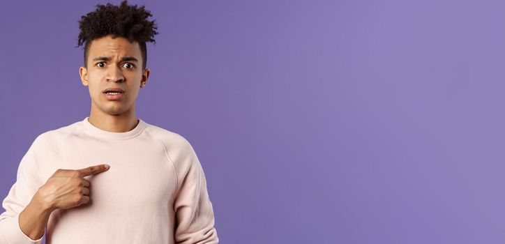 Close-up portrait of embarrassed and worried young man being named or called to principal office, pointing at himself and asking why with disbelief and anxiety, standing purple background.