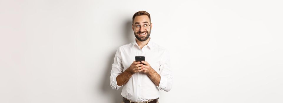 Thouthful handsome manager using mobile phone and thinking about answering message, looking at upper left corner and smiling, standing over white background.