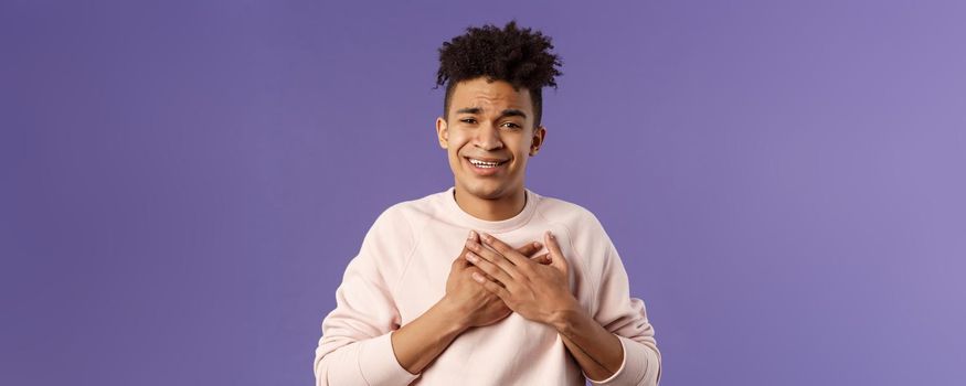Close-up portrait of handsome silly hispanic guy with dreads, place hands on heart and sighing as contemplate something adorable speaking from all his soul, thanking for praises, purple background.