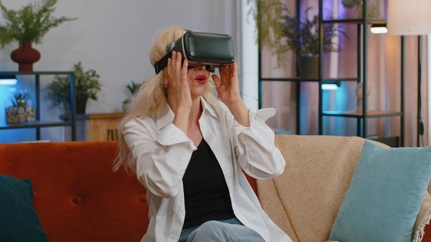Senior old grandmother using virtual reality futuristic technology VR app headset helmet to play simulation 3D video game watching film movie at modern home apartment. Woman in goggles sitting on sofa