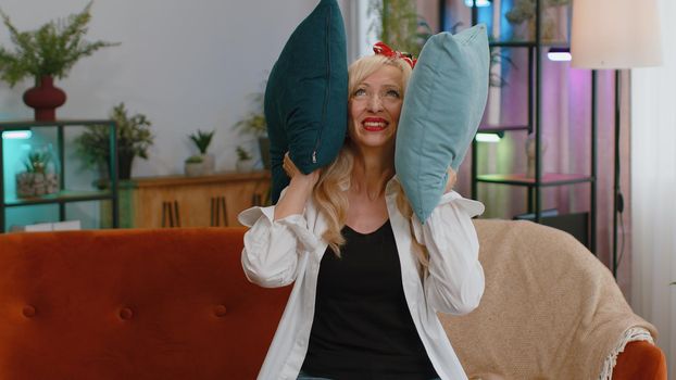 Repair work at neighbours. Irritated senior woman sit on couch cover ears with pillows annoyed by noisy neighbors suffer from headache wishes silence. Thin walls at home flat without sound insulation
