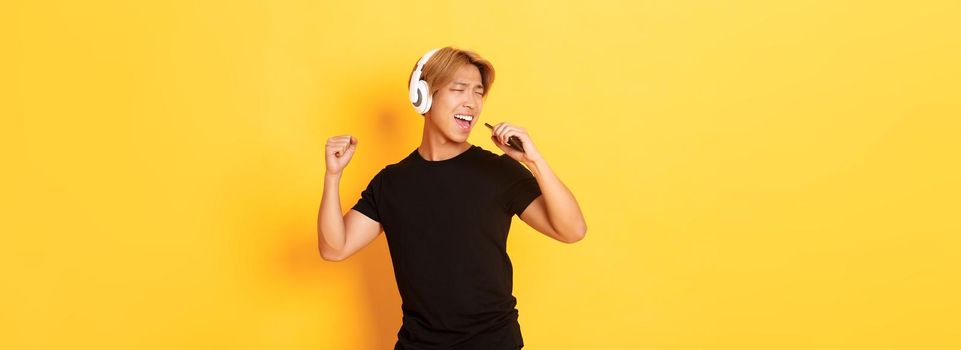 Carefree handsome asian guy in headphones, playing karaoke app, singing into mobile phone microphone, standing yellow background.