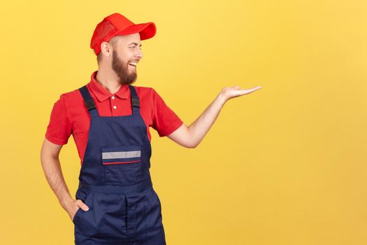 Portrait of smiling worker man standing and presenting copy space for advertisement on his palm, looking away, wearing overalls and red cap Indoor studio shot isolated on yellow background.