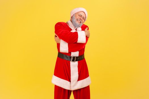 Elderly man with gray beard wearing santa claus costume hugging himself standing with expression of great ego, pleasure and self-esteem. Indoor studio shot isolated on yellow background.