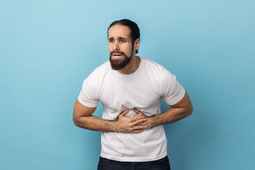 Portrait of ill sick man with beard wearing white T-shirt grimacing and suffering from pain in belly, severe abdominal distress Indoor studio shot isolated on blue background.