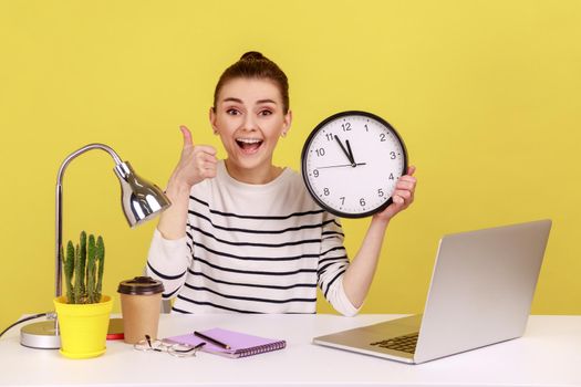 Satisfied happy woman office worker showing thumbs up holding big wall clock, looking at camera with toothy smile, time management. Indoor studio studio shot isolated on yellow background.