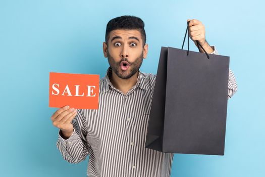Surprised man posing with shopping bags and card with sale inscription in hands, looking at camera with shocked expression, wearing striped shirt. Indoor studio shot isolated on blue background.