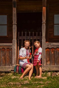 mother and daughter in Ukrainian national costumes are sitting near an old house