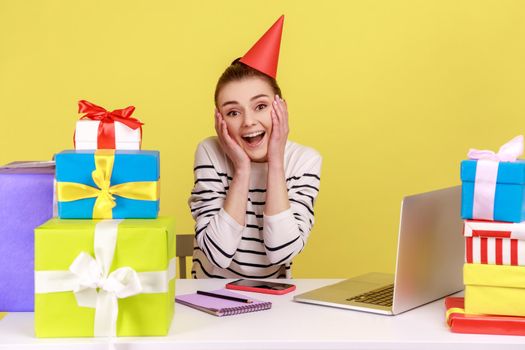 Excited amazed woman secretary sitting on workplace among present boxes, wearing party cone, celebrating her birthday at work. Indoor studio studio shot isolated on yellow background.