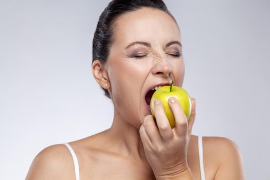 Closeup portrait of middle aged woman biting fresh green apple, uses natural cosmetic, healthy eating, vitamins for health and skin care. Indoor studio shot isolated on gray background.